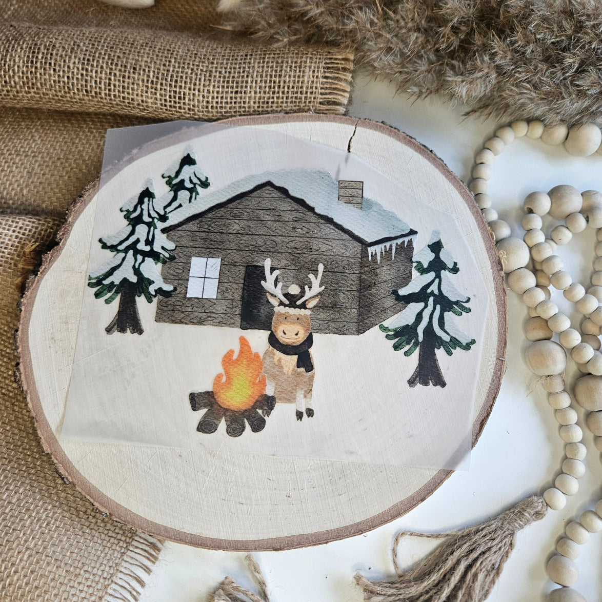 "Rentier Hütte" Bügelbild zum Aufbügeln – niedliches Rentier am Lagerfeuer vor verschneiter Holzhütte mit Tannenbäumen im Aquarellstil auf Holzscheibe, handmade | Little Falili
