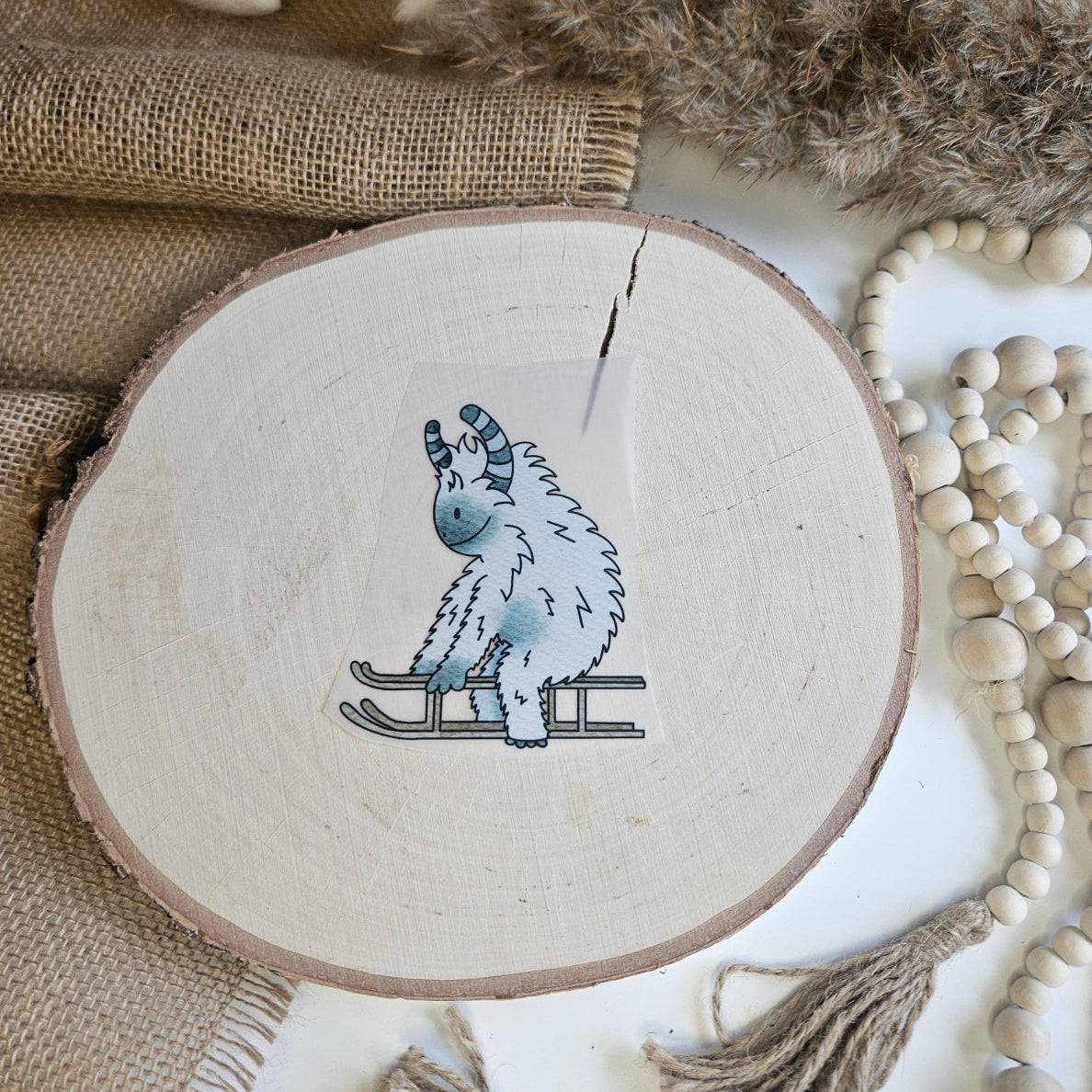 "Winter Yeti" Bügelbild zum Aufbügeln – niedlicher weißer Yeti mit gestreiften Hörnern auf einem Schlitten im Aquarellstil auf Holzscheibe, handmade | Little Falili
