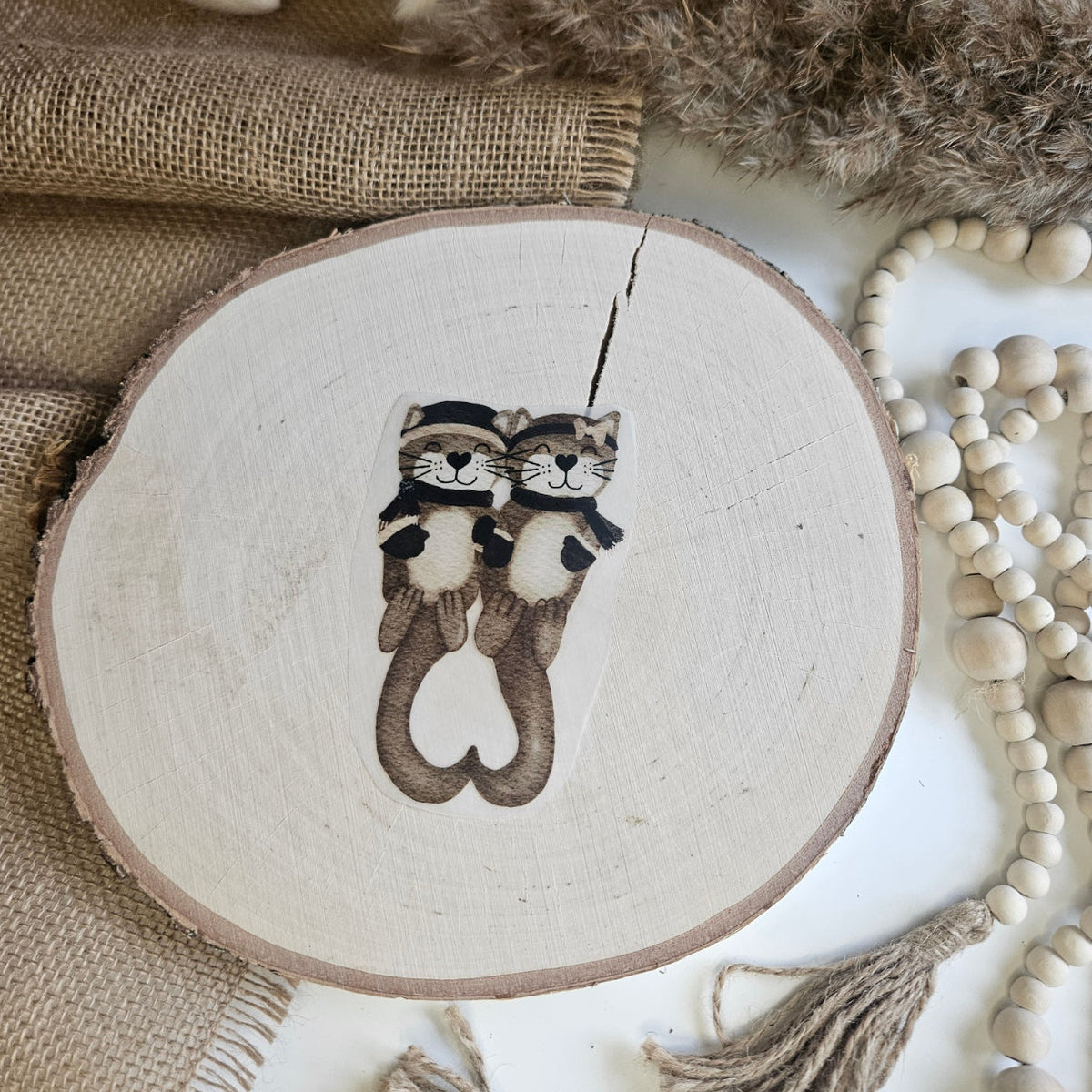 "Winter Kuschelotter" Bügelbild zum Aufbügeln – zwei kuschelnde Otter mit Wintermützen, die ein Herz formen, im Aquarellstil auf Holzscheibe, handmade | Little Falili
