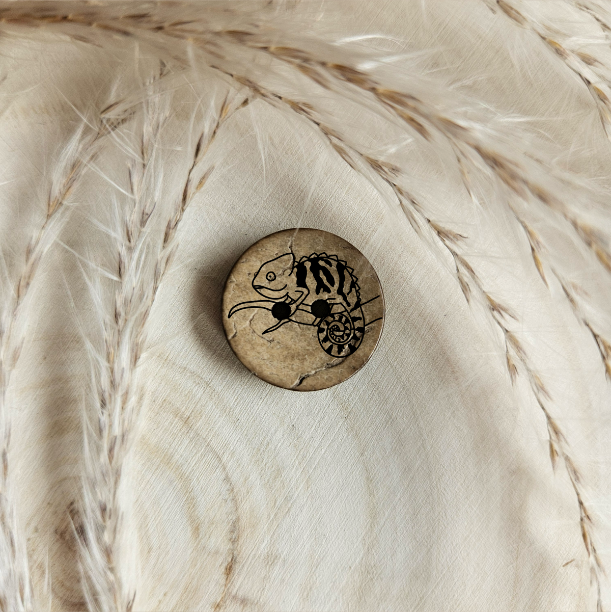 Runder Kokosknopf mit Charmäleonmotiv auf einer hellen Holzscheibe im natürlichen Boho-Stil.