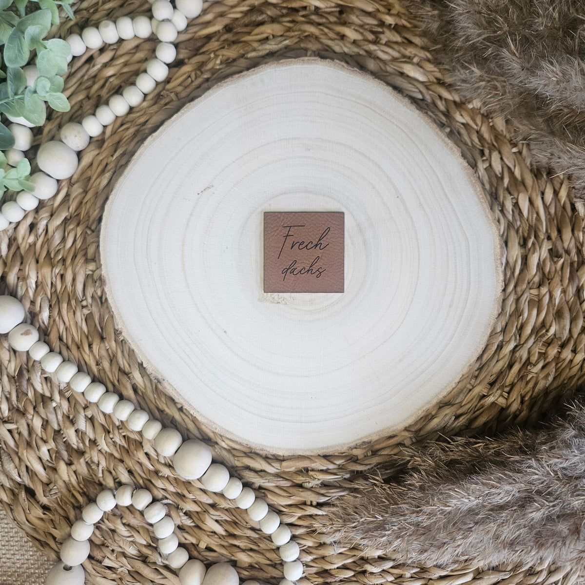 Braunes PU-Kunstlederlabel mit dem Schriftzug „Frechdachs“ auf einer hellen Holzscheibe im natürlichen Boho-Stil.