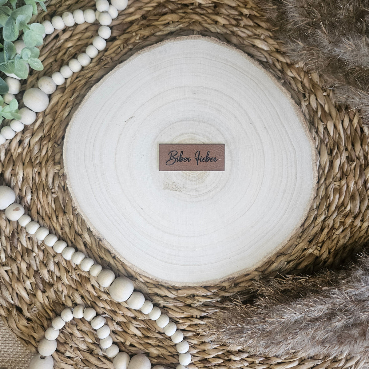 Braunes PU-Kunstlederlabel mit dem Schriftzug „Biber Fieber“ auf einer hellen Holzscheibe im natürlichen Boho-Stil.