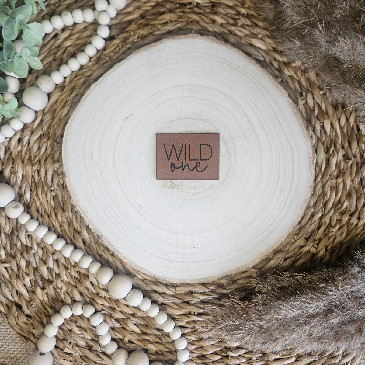 Braunes PU-Kunstlederlabel mit dem Schriftzug „Wild one“ auf einer hellen Holzscheibe im natürlichen Boho-Stil.