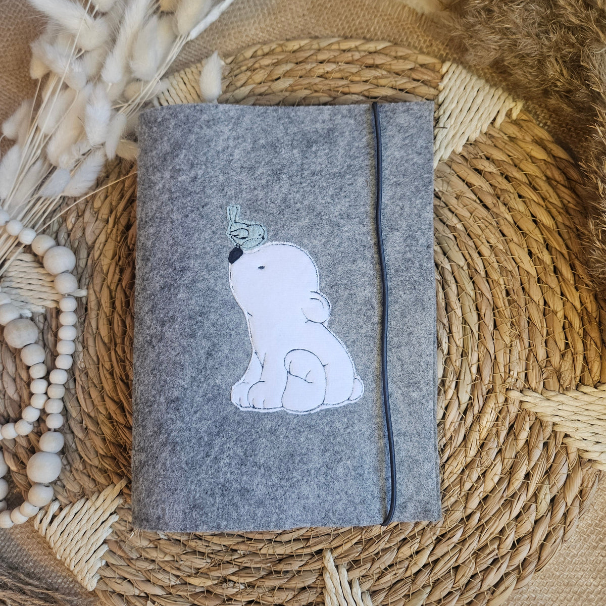 Graue U-Heft-Hülle aus Filz mit Eisbär-Applikation und schwarzem Gummiband auf natürlichem Boho-Hintergrund